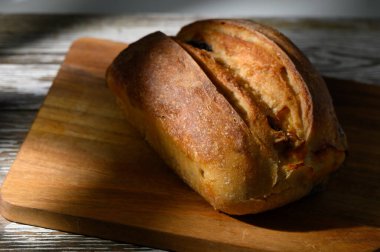 Sıcak bir somun zanaat ekmeği, altın rengi kahverengi ve kabuklu, tahta bir kesme tahtasının üzerinde oturur. Yumuşak ışık davetkar gölgeler yaratır, iyi kullanılmış bir mutfağın ev atmosferini güçlendirir..