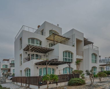 Şık beyaz villa bulutlu bir gökyüzüne karşı duruyor, balkonları ve bereketli yeşillikleri sakin bir kıyı ortamında.