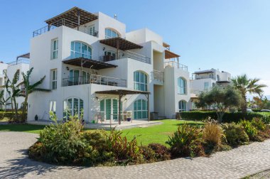 Zarif bir sahil villası, berrak mavi gökyüzünün altında canlı bahçeler ve palmiye ağaçları ile çevrilidir..