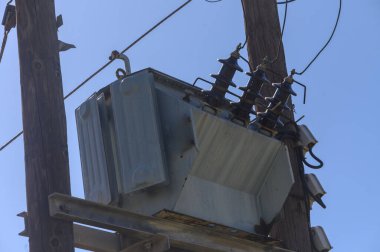 Metalik trafo, uçsuz bucaksız mavi gökyüzüne uzanan kablolarla birlikte tahta bir direğe tünemişti. Kentsel altyapının önemli bir unsuru arka planda sessizce çalışıyor..