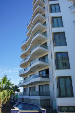 Çarpıcı modern bir binada, berrak bir arka plan ve sallanan palmiye ağaçları karşısında eğimli balkonlar yer alıyor..