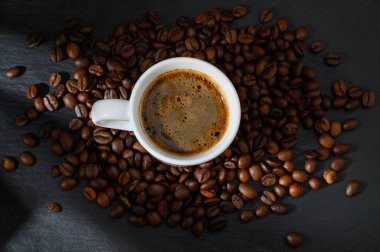 Bir fincan aromatik kahve, koyu kavrulmuş fasulyelerden oluşan bir yatağın üzerinde oturur. Sıcak ve rahat bir ortamda bulunmaya davet eder..