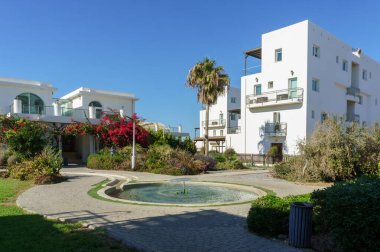 Sakin bir avluda kaynayan bir çeşme, canlı çiçekler ve güneşte güneşlenen beyaz binalar bulunur..