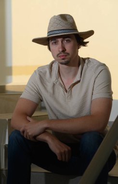 Hasır şapkalı genç bir adam merdivenlerde oturur, rahat bir yaz havası yayar. Sıcak güneş ışığı, öğleden sonranın tadını çıkarırken gevşemiş davranışlarını vurguluyor..