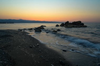 Güneş ufkun altına batarken, canlı renkler gökyüzüne yayıldı. Yumuşak dalgalar kumlu sahili öperken kayalar nöbet tutuyor ve alacakaranlıkta sakin bir atmosfer yaratıyor..