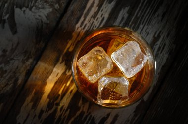 Kehribar sıvısıyla dolu bir bardak ve buz küpleri kırsal bir ahşabın üzerinde duruyor ve akşam ışığının sıcaklığını yakalıyor..