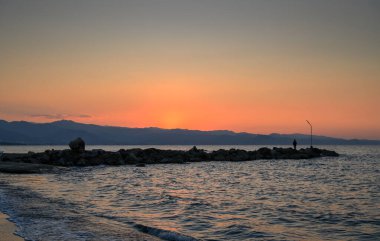 Güneş uzak dağların ardında batarken, sakin kıyı şeridinin huzurlu güzelliğinin tadını çıkaran yalnız bir figür..