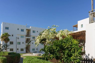 Parlak çevre, açık mavi gökyüzünün altında verimli bahçelerle harmanlanmış beyaz mimariyi sergiliyor..