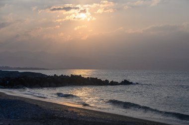 Güneş uzak dağların ardında batarken, altın ışık yumuşak dalgalardan yansıyarak, alacakaranlıkta sakin kıyı şeridi boyunca huzurlu bir ortam yaratır..