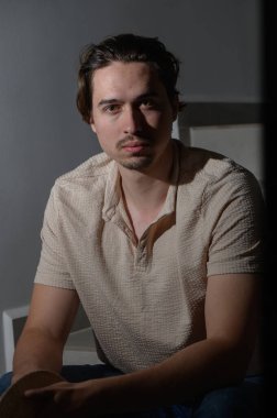 Sakin bir iç mekan ortamında, genç bir adam nazik bir ışık altında düşünceli bir pozda oturur, düşünceli yüz ifadesini ve rahat tavırlarını göstererek gündelik kıyafetlerini giyer..
