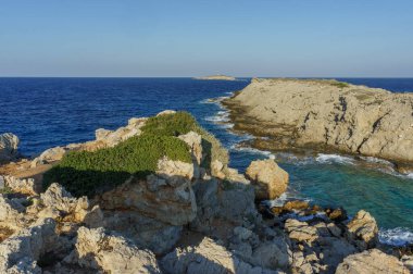 Dalgalar yavaşça yeşil çalılarla süslenmiş kayalık kayalıklara çarpar, sakin bir sahil inzivası sergilerler..
