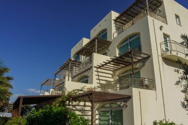 Modern bir bina zarif balkonlara ve gür yeşilliklere sahiptir, canlı mavi gökyüzüne karşı kurulmuştur..