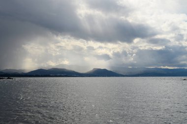 Kara bulutlar tepemizde belirdikçe gölde yumuşak dalgalar dans ediyor. Uzak dağlara gölge düşürüyor. Hava sakin ama potansiyelle dolu. Bu da bir anlık sessizliği çağrıştırıyor..