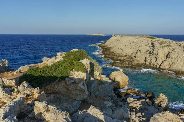 Dalgalar yavaşça yeşil çalılarla süslenmiş kayalık kayalıklara çarpar, sakin bir sahil inzivası sergilerler..