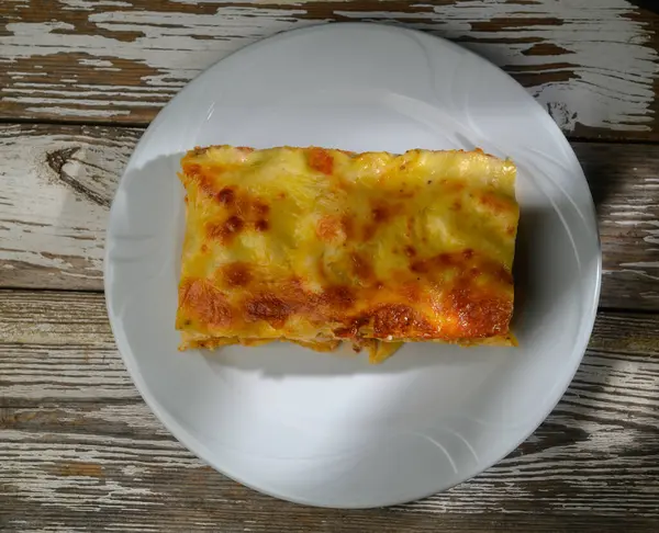 Yeni pişmiş lazanya dilimi peynirle parlıyor, klasik beyaz bir tabakta eskimiş ahşap bir masaya karşı dinleniyor..