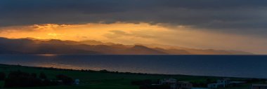 Güneş karanlık bulutların ardında batarken, altın ışınlar sakin denizi ve uzak dağları aydınlatıyor, akşamın soluk ışığında sakin bir atmosfer yaratıyor..