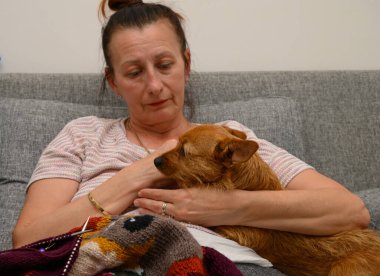 Yumuşak gri bir koltukta oturan bir kadın küçük köpeğini nazikçe kollarına alıyor. Sessiz bir anı paylaşıyorlar, battaniyeye sarılıyorlar, rahat bir akşamda rahatlıklarını ve bağlarını sergiliyorlar..