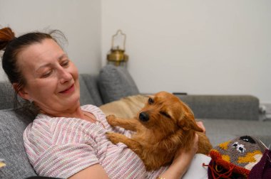 Bir kadın rahat bir kanepeye oturur, küçük köpeğini kucağına yatırır. Huzurlu bir yaşam alanıyla çevrili, akşam ışığının yumuşak parıltısında keyifli bir anı paylaşıyorlar..