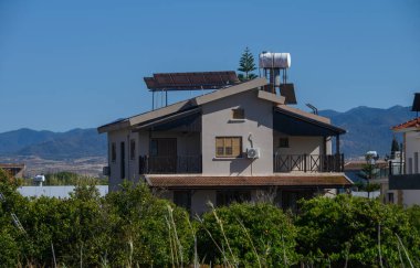 Huzurlu bir manzarada, çağdaş bir ev yemyeşil ile çevrili duruyor. Güneş ışığı, berrak mavi gökyüzünün altındaki görkemli dağların arka planıyla, sahnenin canlı renklerini güçlendirir..