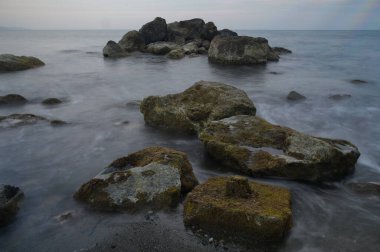 Yumuşak dalgalar sert kayalara çarpar, yüzeyleri canlı yosunlarla süslenir. Batan güneş, bu huzurlu plajın üzerine yumuşak bir parıltı saçar. Sessiz bir yansıma ve güzellik anı yaratır..