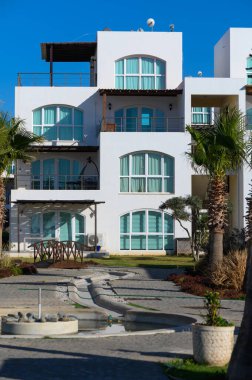 Bereketli palmiye ağaçları, geniş pencereli, sakin sulara bakan modern, beyaz duvarlı yapıları çerçevelemekte ve güneş ışığıyla yıkanmış bir sahil inzivasında rahatlamaya davetiye çıkarmaktadır..