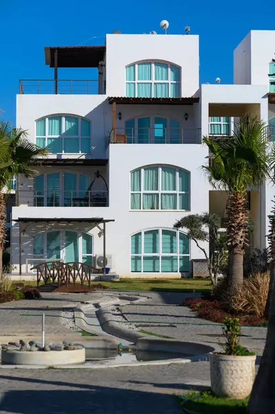 Bereketli palmiye ağaçları, geniş pencereli, sakin sulara bakan modern, beyaz duvarlı yapıları çerçevelemekte ve güneş ışığıyla yıkanmış bir sahil inzivasında rahatlamaya davetiye çıkarmaktadır..