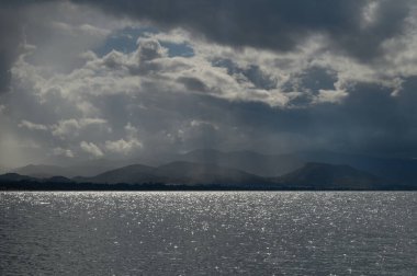 Akşam yaklaşırken, karanlık bulutlar, titreyen ışığı yansıtan sakin suların üzerinde kaygı verici bir şekilde toplanır. Uzak dağlar, değişen gökyüzünün altında dramatik bir zemin hazırlıyor..