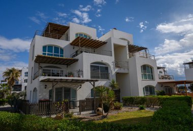 Modern tasarıma sahip şık beyaz bina, yeşil ve mavi gökyüzü ile çevrili, sakin bir sahil havası yakalıyor..