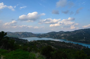 Parlak bulutlar berrak tepelerle çevrili sakin bir göl üzerinde canlı mavi bir gökyüzü boyunca sürükleniyor. Doğa, sıcak güneşin altında huzur içinde, dinlenmeyi ve tefekküre yol açar..