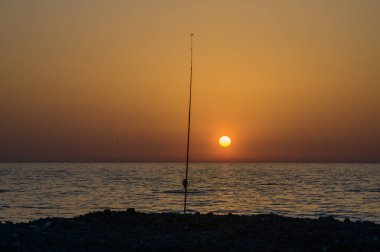 Sakin su, akşam gökyüzünün ılık tonlarını yansıtır. Balık tutmak için mükemmel sakin bir kıyı atmosferi yaratır..