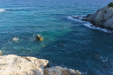 Dalgalar, sakin mavi sularda, huzurlu bir ortamda güneş ışığını yansıtan engebeli kıyı şeridini hafifçe okşar..