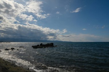 Güneş ışığı yukarıdaki dağınık bulutların arasından geçerken dalgalar yavaşça kıyıdaki kayalara çarpıyor. Uzakta dağlar belirir, sakin ve güzel bir manzarayı işaret eder..