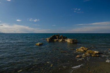 Dalgalar, turkuaz sudan gün ışığına çıkan bir kaya kümesi gibi kıyı şeridine hafifçe çarpar. Uçsuz bucaksız gökyüzü yumuşak bulutlarla kaplı, sakin bir atmosfer yaratıyor..