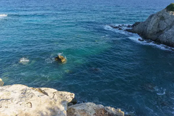 Dalgalar, sakin mavi sularda, huzurlu bir ortamda güneş ışığını yansıtan engebeli kıyı şeridini hafifçe okşar..