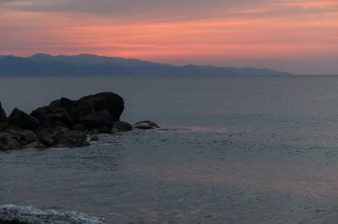 Güneş ufukta batarken yumuşak dalgalar kayalara çarpar, gökyüzünü pembe ve mor renklerle boyarlar. Sakin bir atmosfer, alacakaranlıkta sahil güzelliğini arttırır..