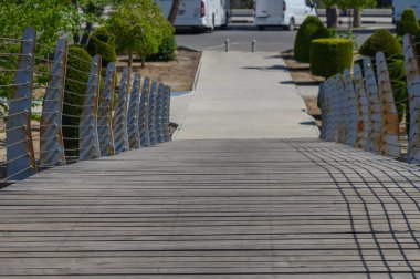 Büyüleyici ahşap bir köprü size huzur dolu bir parkta yol gösterir. Çitleri budanmış ve güneşli beton bir yolda keyifli bir gezinti için..