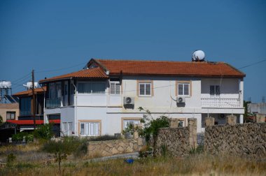 Kırmızı kiremit çatılı ve birden fazla penceresi olan iki katlı tuhaf bir ev, parlak mavi gökyüzünün altında sakin bir kırsal alana yerleşmiş. Çevreleyen yeşillik sükuneti artırır.