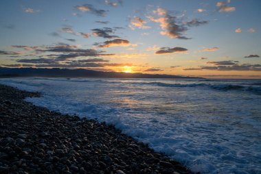 Güneş ufukta batarken, kayalık kıyı şeridinde sıcak bir parıltı oluşturan altın renkler gökyüzünü dolduruyor. Dalgalar taşlara ritmik olarak çarpar ve sakin bir atmosfer yaratır..