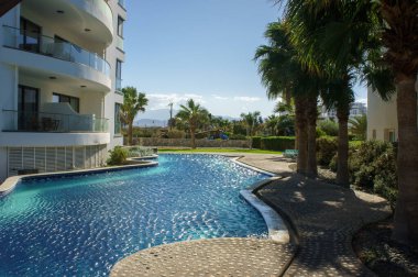 Sersemletici mavi bir havuzu ve palmiye ağaçları olan sakin bir vaha, güneşli bir gökyüzü altında rahatlamaya davetiye çıkarıyor..