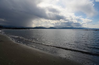 Huysuz bulutlar sakin suyun üzerine gölgeler düşürüyor, sakin bir ortam yaratıyor, alacakaranlıkta yumuşak dalgalar kıyıya vuruyor, huzur ve tefekkür hissi uyandırıyor..