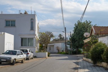 Huzurlu bir yerleşim bölgesinde beyaz evler, yeşil ağaçlar ve güneşli mavi gökyüzünün altında park edilmiş arabalar yer almaktadır..