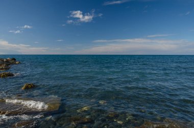 Yumuşak dalgalar kayalık kıyı şeridinde yuvarlanır, parlak mavi gökyüzünde kayan tüylü bulutlar gibi güneş ışığını yansıtır. Huzurlu atmosfer, deniz kenarında huzurlu bir tefekküre davetiye çıkarır..