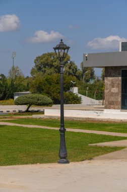 Eski tarz bir sokak lambası, yeşil ağaçlar ve çağdaş bir yapı tarafından çerçevelenmiş güneş ışığı parkına biraz zarafet katıyor. Sahne huzurlu ve davetkar bir atmosfer yayıyor..