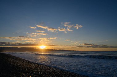 Gün ışığı söndükçe, güneş ufkun altına batar, gökyüzünü turuncu ve altın rengine boyarken, nazik dalgalar bulutlu bir gökyüzünün altında kumlu sahili öper..