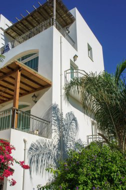 Güzel beyaz bir villa canlı yeşilliklerin ortasında zarif bir şekilde duruyor, bir kıyı inzivasının sıcak güneşinin altında güneşleniyor..