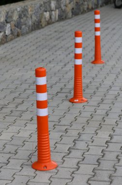 Üç canlı turuncu trafik konisi iyi döşenmiş kaldırım taşı bir yolda dikiliyor ve trafik için bariyer oluşturuyor. Kalın çizgileri dikkati çeker, şehir manzarasında hareketi yönetir..