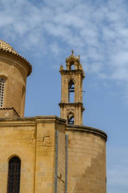 Tarihi bir kilisenin çarpıcı manzarası ayrıntılı çan kulesini ve pürüzsüz taş duvarlarını sergiliyor, hepsi dağınık bulutlarla süslenmiş parlak mavi bir gökyüzünün altında..