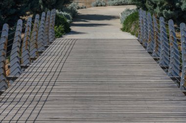 Yıpranmış ahşap bir köprü, gezginleri berrak bir öğleden sonra güneşin yansıttığı canlı yeşillik ve gölgelerle çerçevelenmiş sakin bir patikanın karşısına davet eder..