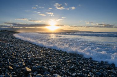 Altın ışınlar kıyı boyunca çakıl taşlarını aydınlatırken dalgalar alacakaranlıkta canlı bir gökyüzü altında yavaşça çarpışıyor..