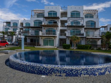 Parlak beyaz dairelerde berrak mavi bir gökyüzünün altında sakin dairesel bir havuz çevreleyen balkonlar yer alıyor..